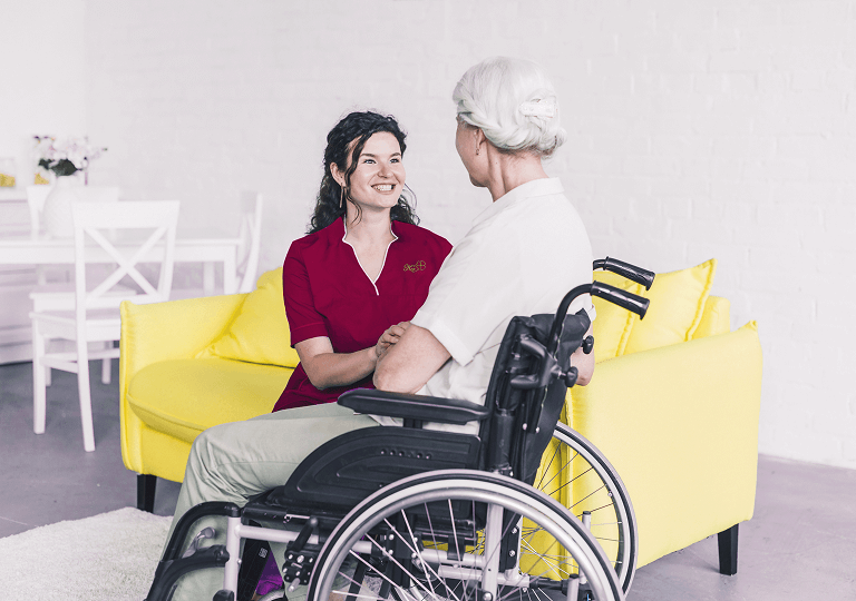 Pflegekraft im roten Outfit sitzt auf einem gelben Sofa und spricht einfühlsam mit einer älteren Frau im Rollstuhl in einem hellen, freundlich eingerichteten Raum.
